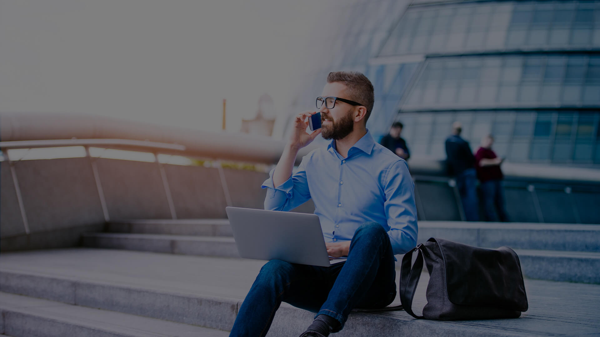 Man in business casual attire talking on the phone and sitting on steps outside with a messenger bag next to him 