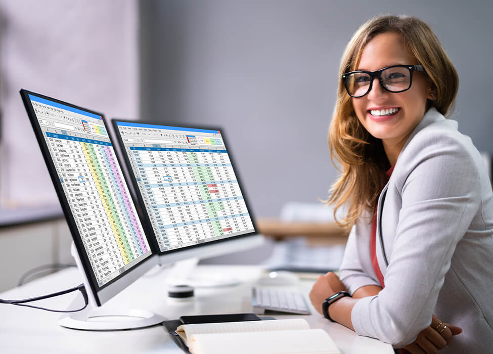 Smiling female healthcare professional at a desk with dual monitors displaying extensive medical coding analytics, demonstrating RVU financial optimization and medical billing accuracy
