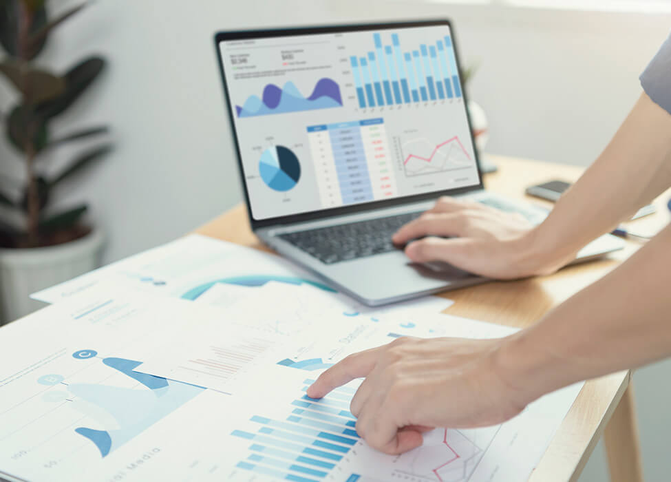 Hands pointing to charts and graphs on a laptop screen and printed documents, representing detailed data analysis and real-time data insights for strategic decision-making.