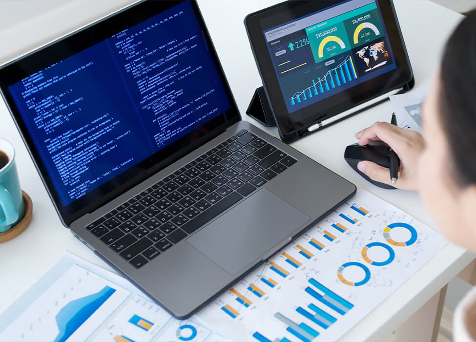 A professional working on a laptop displaying lines of code and real-time data analysis charts on a tablet. The desk is cluttered with printed charts and graphs, indicating an in-depth analysis of data trends.