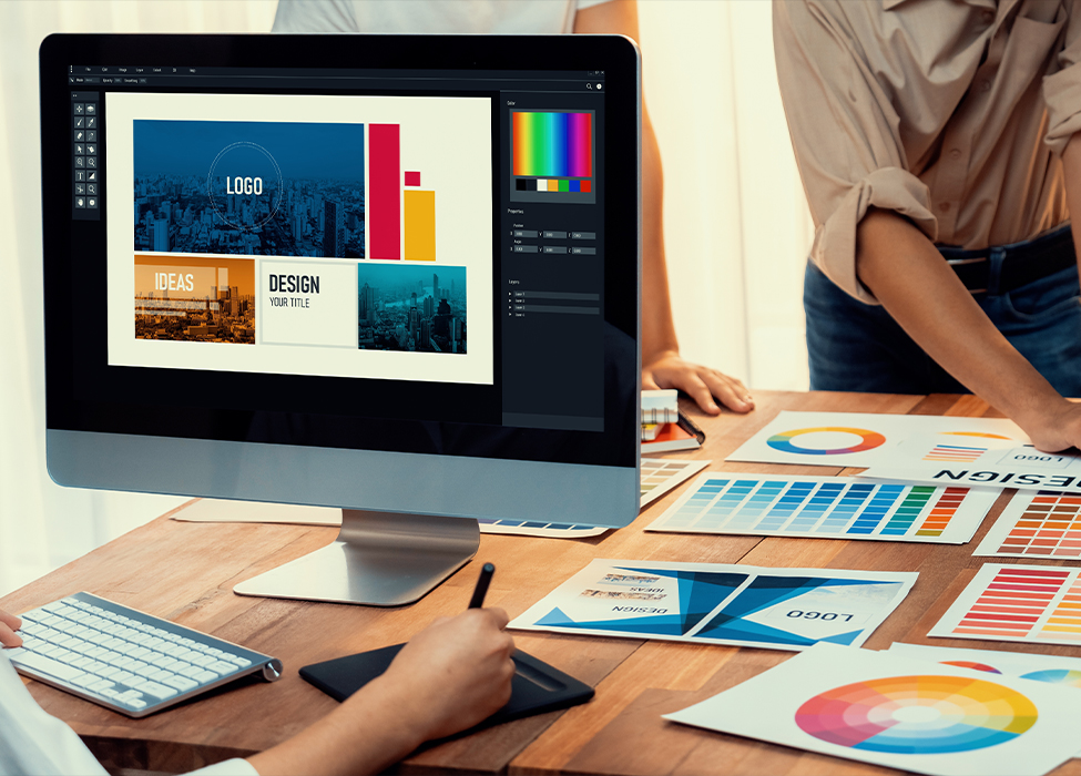 Image of a monitor with graphics currently being worked on while two other people are standing next to the table looking at papers