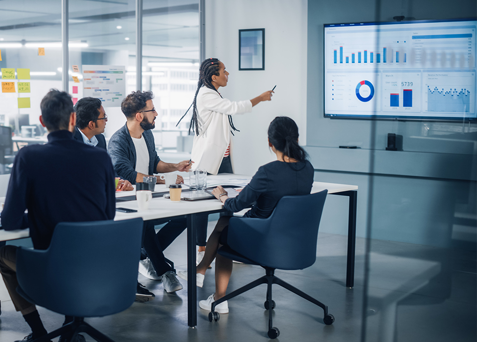 People sitting around a table having a meeting while looking at a screen with graphs on it