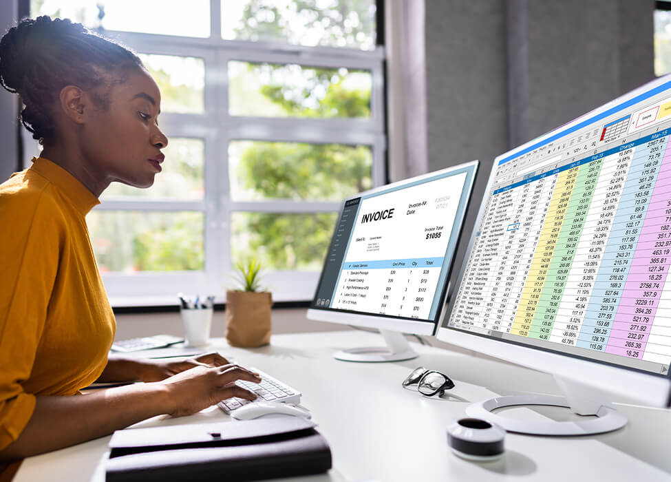 A professional working on a dual-monitor setup, with one screen displaying a detailed invoice and the other showing a spreadsheet with various data points. This could be related to a medical coding audit where financial and medical records are analyzed.