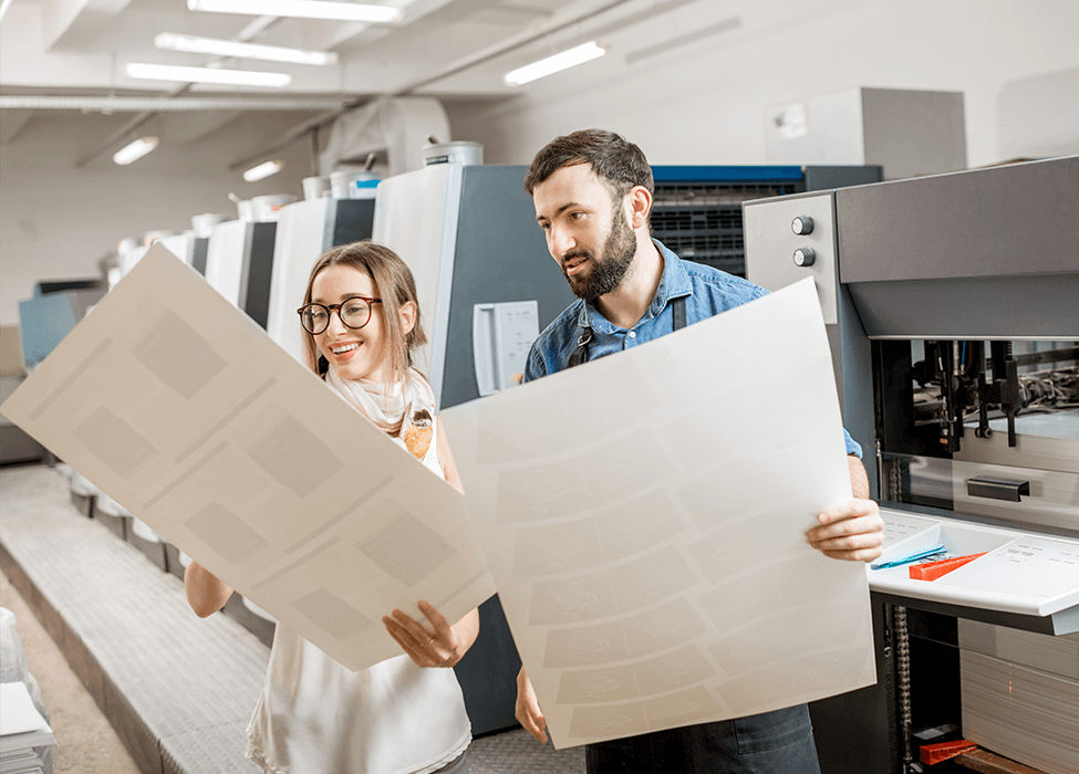 Two colleagues, a man and a woman, smiling while examining large printed sheets in a print shop, showcasing the collaboration aspect of managed print services.