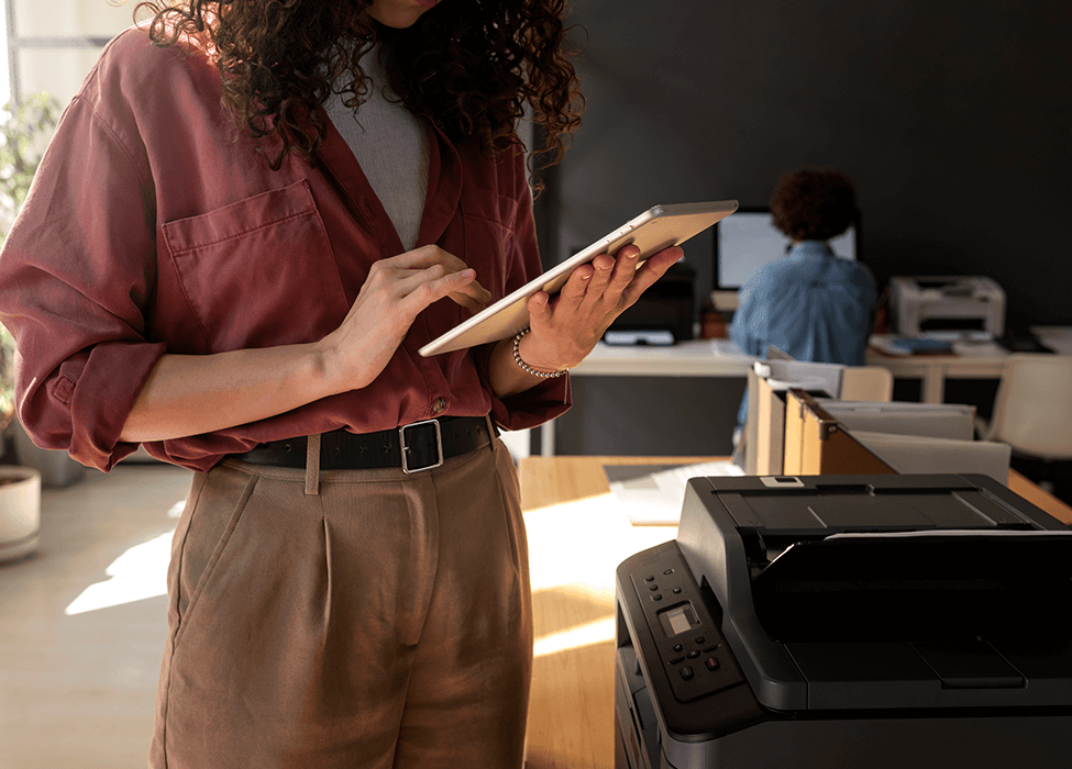 A young woman using a digital tablet while standing next to a printer in a bright office environment, symbolizing efficient managed print services.