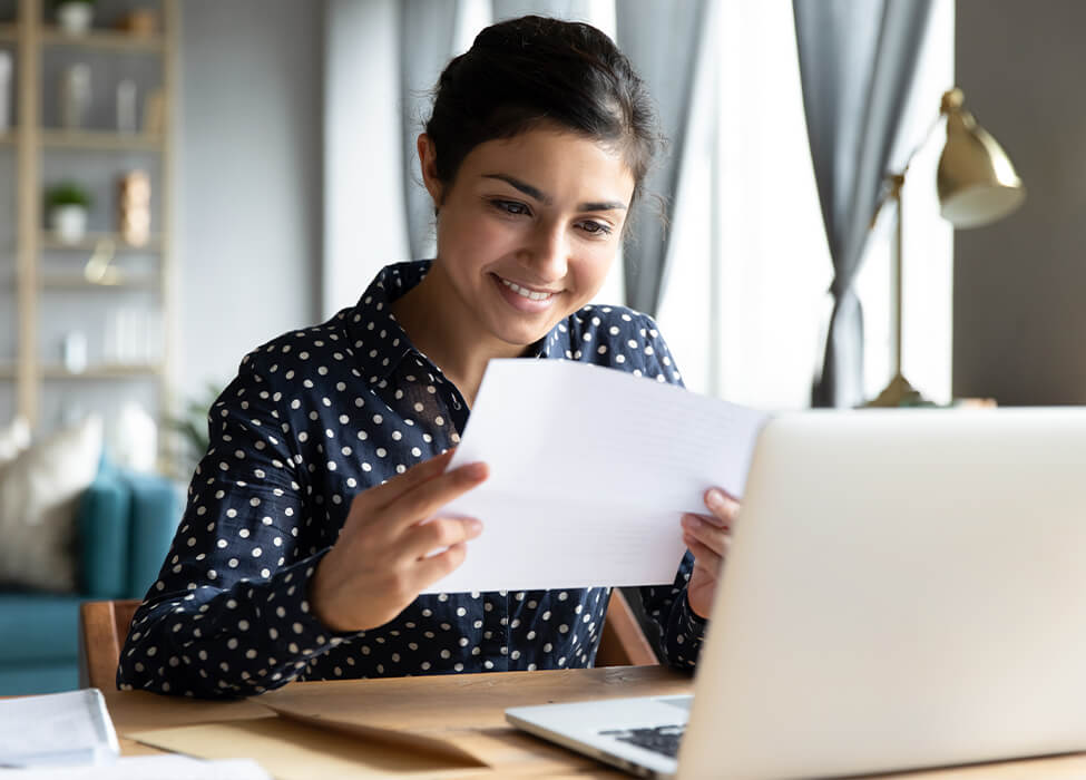 Woman sitting at desk with laptop, smiling at the paper she's holding
